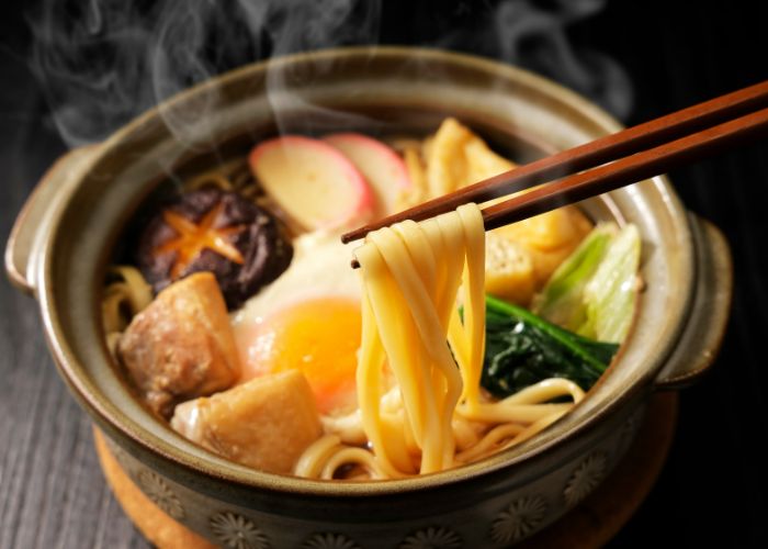 A steaming pot of nabeyaki udon, including udon noodles, mushrooms, fishcakes, and a boiling egg.